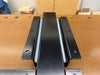 A black metal woodworking vise mounting bracket on a wooden workbench, with a light blue panel visible behind them.