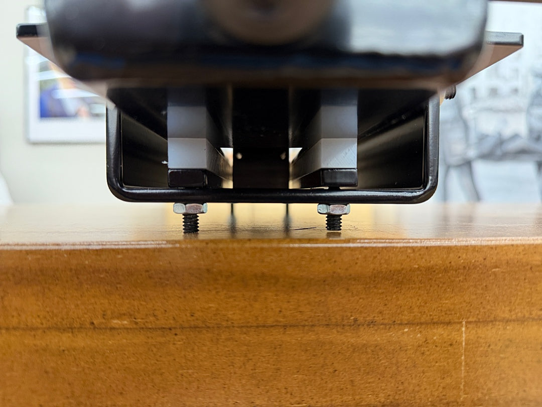 Close-up of a black metal mounting bracket with two visible screws fastened onto a wooden surface, with a blurred indoor background.