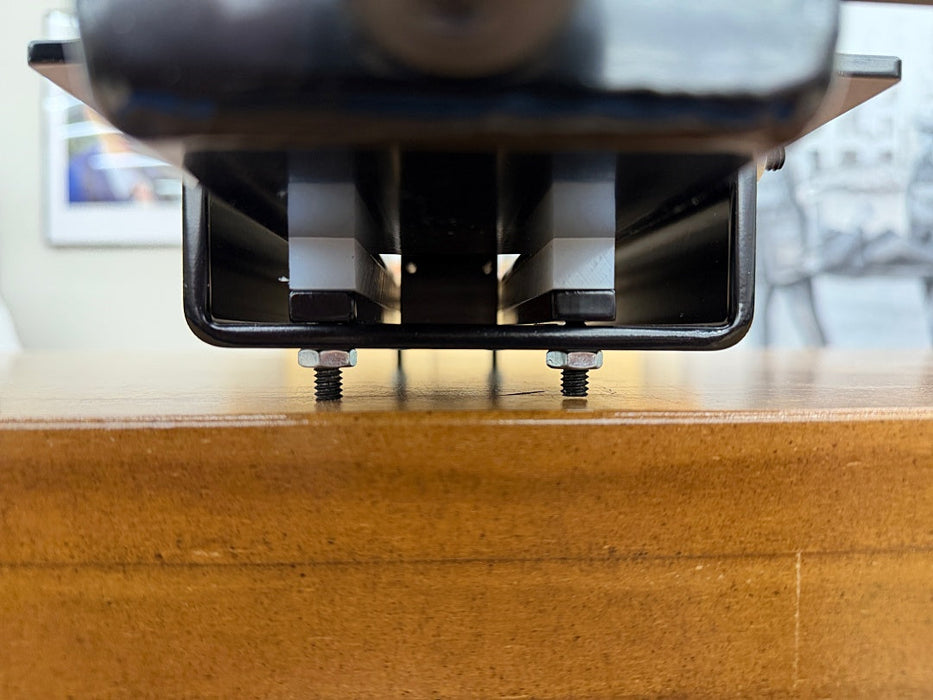 Close-up of a black metal mounting bracket with two visible screws fastened onto a wooden surface, with a blurred indoor background.