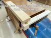 A close-up of a wooden bench vise with a brass adjustment nut and a wooden handle, mounted on a wooden workbench in a workshop. White text overlay reads 'Vise Jaw and Brass Bench Dog Sold Separately'.