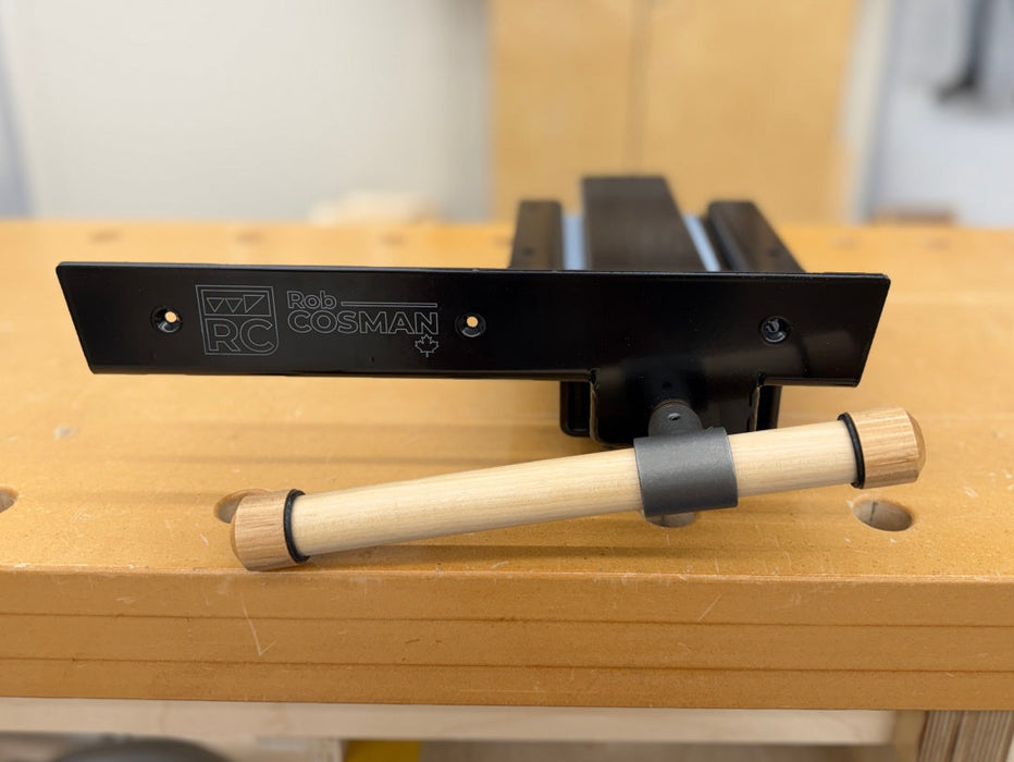 A close-up of a metal Rob Cosman Workbench Vise resting on a wooden workbench with a wooden-handled tin front.